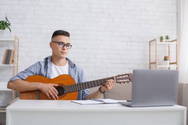 Covid-19 salgını sırasında evde modern hobi ve eğitim. Gözlüklü erkek öğrenci gitar çalıyor ve oturma odasında dizüstü bilgisayara bakıyor.