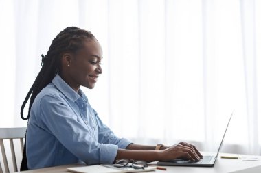 Gülümseyen Afrikalı Serbest Kadın Laptop 'ta çalışıyor, Pencere kenarındaki masada oturuyor