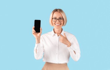 Resmi giyinmiş pozitif kadın sekreter mavi stüdyo arka planında boş ekranlı akıllı telefon gösteriyor.