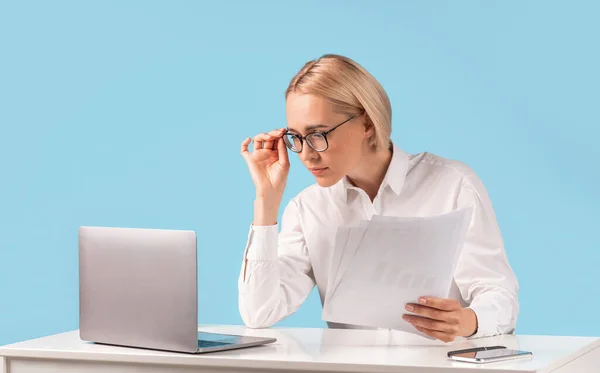 Сотрудница женского офиса просматривает документы, делает отчет, делает бумажную работу рядом с ноутбуком на синем фоне — стоковое фото