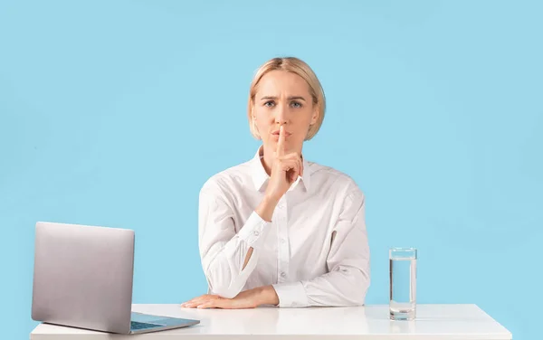 Attraente donna d'affari che fa un gesto di silenzio sul posto di lavoro vicino al computer portatile, sfondo blu studio — Foto Stock