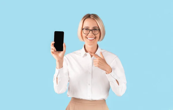 Positive female secretary in formal clothes showing smartphone with empty screen over blue studio background