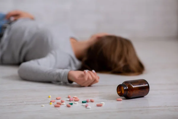 Jonge vrouw die zelfmoord pleegt door een overdosis pillen, focus op de fles tabletten verspreid over de vloer. Depressieconcept — Stockfoto