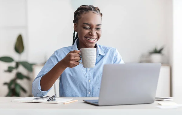Χαρούμενη μαύρη γυναίκα υπάλληλος γραφείου χρησιμοποιώντας φορητό υπολογιστή και πίνοντας καφέ στο χώρο εργασίας — Φωτογραφία Αρχείου