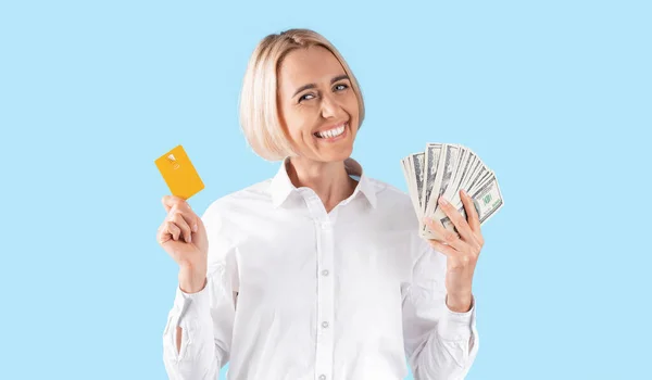 Retrato de una mujer de negocios positiva mostrando dinero y tarjeta de crédito sobre fondo de estudio azul, panorama — Foto de Stock