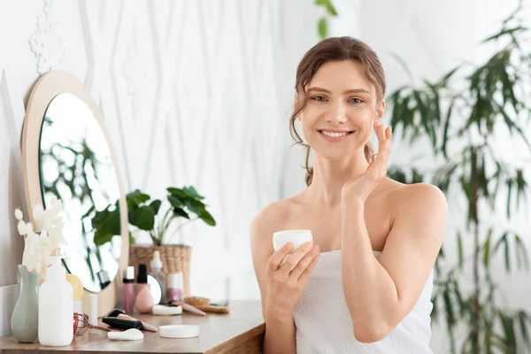 Sorridente giovane signora con crema viso in camera da letto — Foto Stock