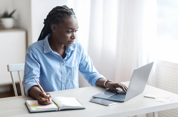 Молодая черная фрилансер женщина работает на ноутбуке в домашнем офисе, делая заметки — стоковое фото