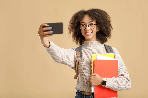 Back to college or university after quarantine and photo for blog. Happy african american lady in glasses, with backpack and notepads makes selfie on smartphone