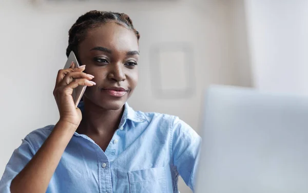 Успішна чорна бізнес-леді говорить на мобільному телефоні і дивиться на екран ноутбука — стокове фото