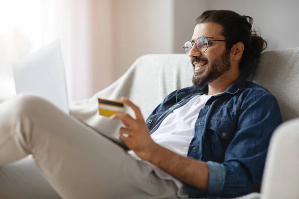 Cheerful Guy Using Mobile Phone, Credit Card And Laptop Providing Online Payment Sitting On Couch At Home.