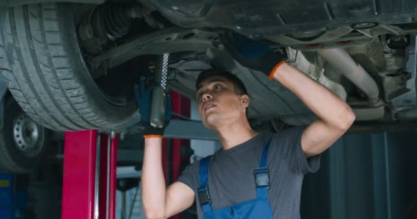 Młody mechanik naprawiający zawieszenie samochodu, stojący pod pojazdem w garażu — Wideo stockowe