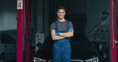 Kirli oto tamircisinin portresi, atölyede arabanın yanında poz vermek, kameraya gülümsemek.