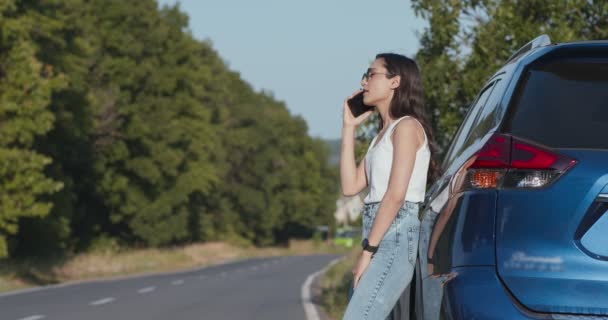 좌절감에 빠진 젊은 여자가 휴대폰으로 말하면서, 자동차에 경고 신호를 보내며 의지하고 있다 — 비디오