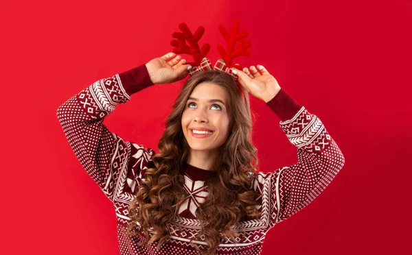 E 'il periodo natalizio. Bella giovane donna in maglione di lana che indossa corna di cervo su sfondo rosso studio, panorama — Foto Stock