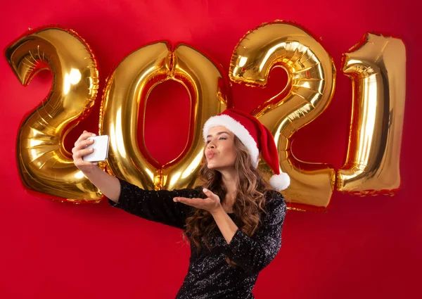 Elégante femme yong dans le chapeau de Père Noël et belle robe de soirée prenant selfie sur la veille du Nouvel An 2021, fond studio rouge — Photo