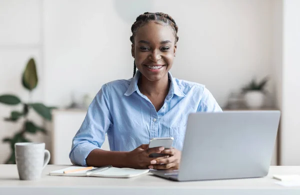 Fiatal fekete nő irodai alkalmazott ül a munkahelyen okostelefonnal a kezében — Stock Fotó