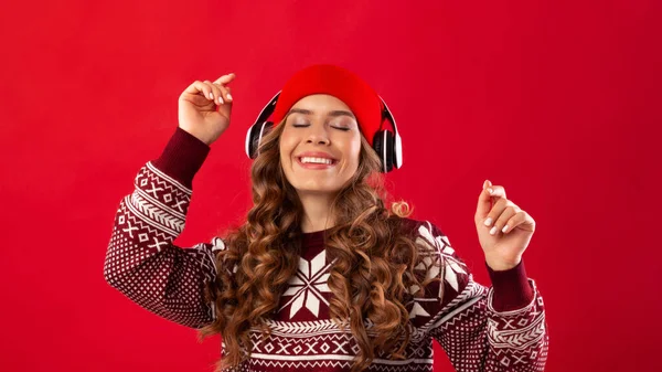 Kerstmuziek. Mooie jonge vrouw in koptelefoon en winter outfit dansen op haar favoriete lied op rode achtergrond — Stockfoto