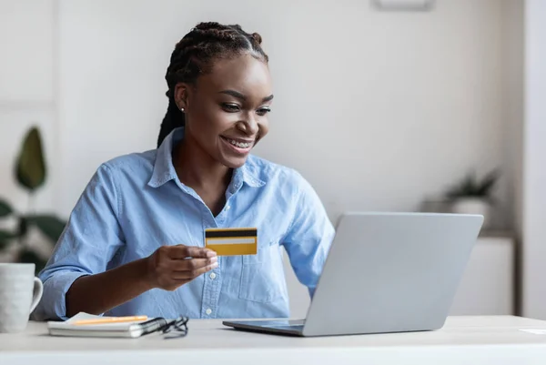 Pago en línea. Negocio negro sonriente usando el ordenador portátil y la tarjeta de crédito en la oficina — Foto de Stock