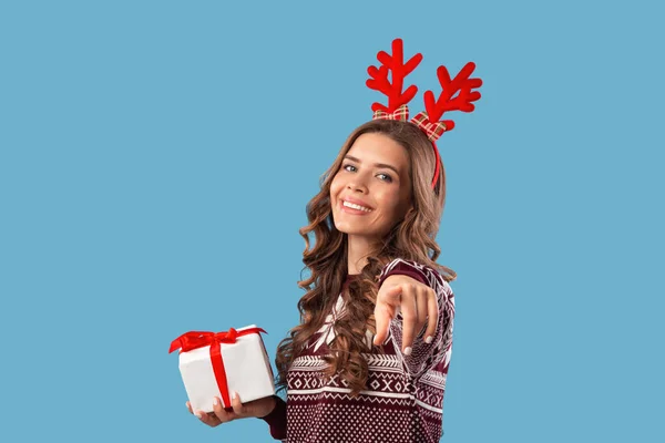 Encantadora dama milenaria en traje de Navidad sosteniendo la caja de regalo y señalando con el dedo a la cámara en el fondo azul del estudio — Foto de Stock