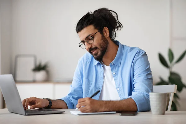 Der westliche Freiberufler Guy macht sich Notizen zum Notizblock, während er im Home Office arbeitet — Stockfoto