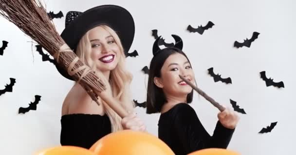 Encantadoras de Halloween. Dos mujeres con disfraces sonriendo y coqueteando a cámara, sujetando escoba y varita mágica — Vídeos de Stock