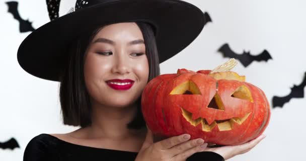 Sly asiatisk kvinna i häxa kostym håller snidade Halloween pumpa och skrattar, vit studio bakgrund med papper fladdermöss — Stockvideo