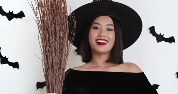 Весела азіатська жінка чарівник в чорному костюмі з мітлою, посміхається на камеру і махає привіт, білий фон — стокове відео