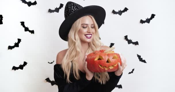 Halloween tradities. Schattige blonde vrouw in heks kostuum poseren met gesneden jack-o-lantaarn pompoen — Stockvideo