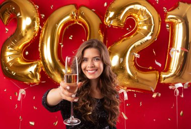 Yeni yıl partisinde bir bardak şampanyayla kadeh kaldıran genç ve güzel bir kadın. Kırmızı stüdyo arka planında altın balonlarla.