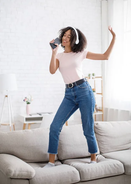 Aplicación móvil musical y micrófono imaginario. Divertida mujer afroamericana bailando en un sofá — Foto de Stock