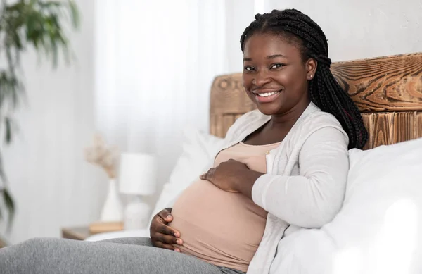 Menikmati Kehamilan. Wanita hamil hitam yang ceria beristirahat di tempat tidur, menyentuh perutnya — Stok Foto