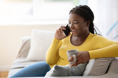 Neşeli siyah kadın cep telefonuyla konuşuyor, kahve içiyor, koltukta dinleniyor.
