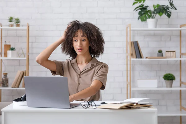 Tarea difícil o problemas en el trabajo en línea. Sorprendida mujer afroamericana se rasca la cabeza y mira el portátil — Foto de Stock