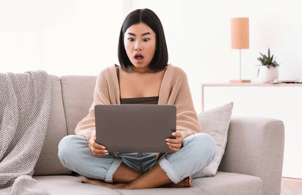 Überraschte asiatische Frau sitzt auf Couch und benutzt Laptop — Stockfoto