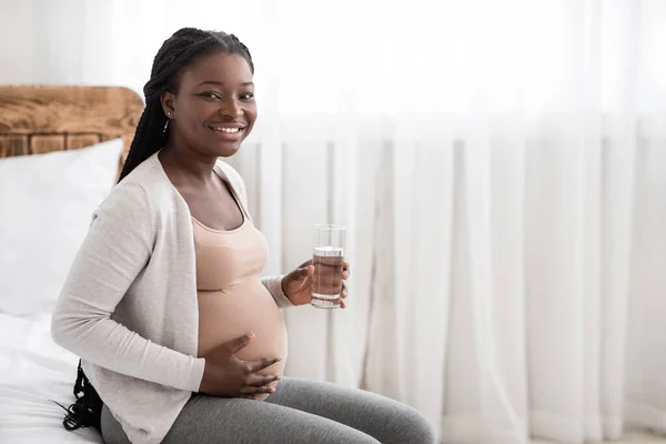 Gelukkig Afrikaans zwanger vrouw drinken water uit glas terwijl ontspannen op bed — Stockfoto