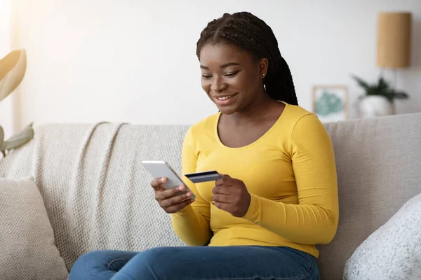 Compras online. Sorrindo preto senhora usando Smartphone e cartão de crédito em casa — Fotografia de Stock