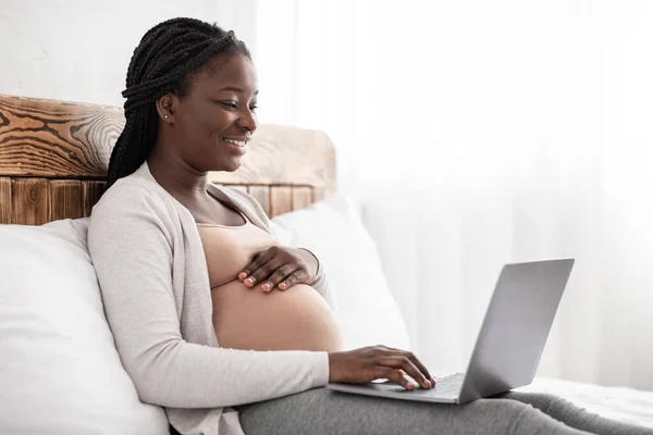 Очаровательная беременная женщина отдыхает с ноутбуком, просматривает или болтает с друзьями онлайн, сидит на кровати, панорама с пустым пространством — стоковое фото