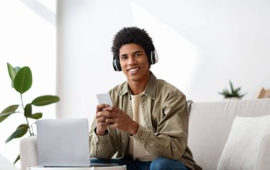 Kulaklığı, bilgisayarı ve cep telefonu olan komik siyahi bir genç evden internetten ders çalışıyor.