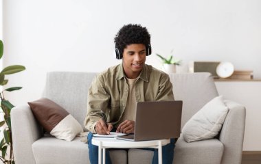 Web tabanlı eğitim konsepti. Kulaklıklı mutlu genç adam dizüstü bilgisayardaki öğretmenle çevrimiçi iletişim kurarken not alıyor.