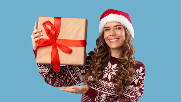 Comprando para Navidad. Dulce mujer milenaria en Santa sombrero mostrando caja de regalo envuelta con cinta sobre fondo azul — Foto de Stock