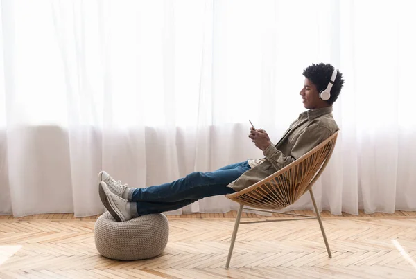 Afro-americano adolescente cara com fones de ouvido ouvir música no smartphone enquanto sentado na cadeira em casa, espaço de cópia — Fotografia de Stock