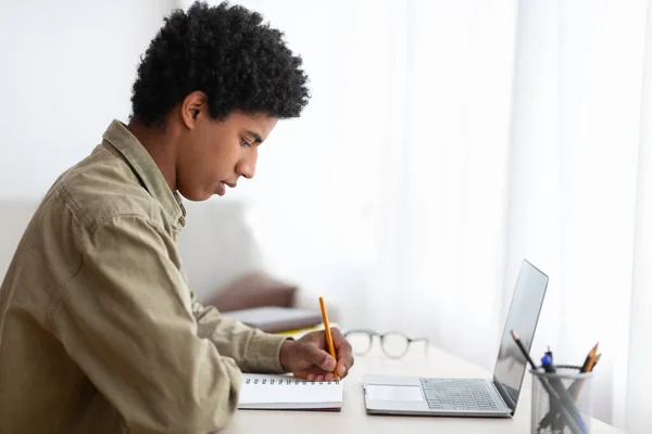 Conceito de educação remota. Visão lateral do cara adolescente preto tomar notas em Copybook durante conferência na web em casa — Fotografia de Stock