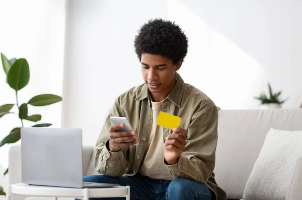 Ragazzo adolescente nero positivo con laptop utilizzando smartphone e carta di credito per lo shopping online a casa — Foto Stock