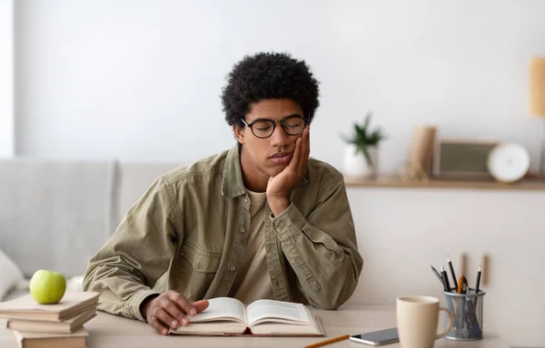 Concepto de educación remota. Cansado estudiante negro durmiendo en la mesa mientras lee el libro de texto en el interior — Foto de Stock