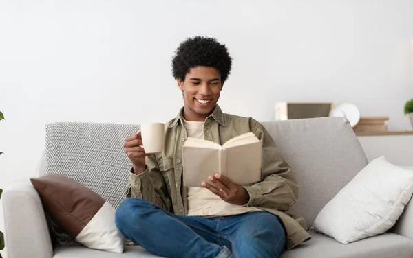 积极的黑人青少年坐在沙发上看书和喝咖啡 — 图库照片