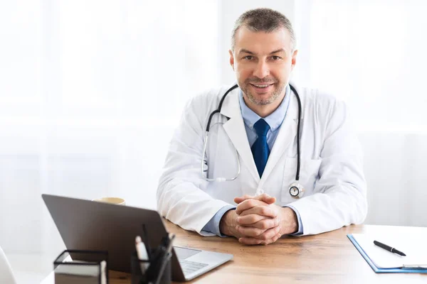 Portrait of mature doctor looking at camera sitting at desk — Stock Photo, Image