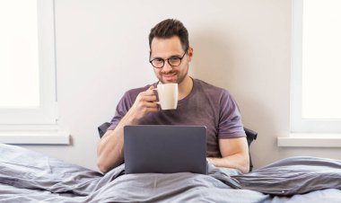 Adam dizüstü bilgisayar kullanıyor, film izliyor, kahve içiyor, yatak odasında oturuyor.