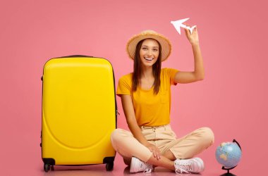 Mutlu bayan kağıt uçakla oynuyor, bavulun yanında oturuyor.