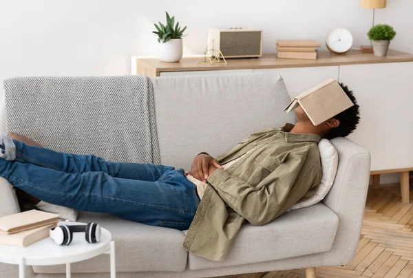 Conceito de educação remota. Entediado adolescente estudante preto com livro em seu rosto dormindo no sofá em casa — Fotografia de Stock