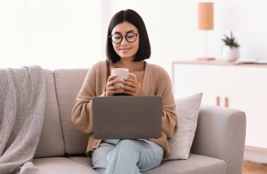 Asyalı kadın bilgisayar kullanıyor ve sıcak kahve içiyor.